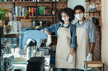 Coffee shop owners with african american male and female in aprons, white gloves and protective face masks in modern interior